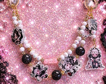 May include: A decorative beaded chain featuring white and black glittery spherical beads, interspersed with gold-toned spacers and clear acrylic charms depicting illustrated characters and the text CHANEL. The chain ends in two gold-toned circular clasps.
