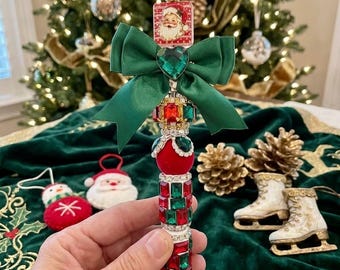 May include: A decorative pen featuring a Santa Claus square topper, a green satin bow, and a barrel encrusted with red and green faceted rhinestones. The pen is accented with a heart-shaped green gem and a white beaded finial at the very top.