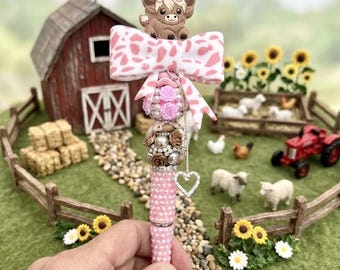 May include: A decorative pen featuring a brown highland cow topper, a pink and white cow-print bow, and a barrel adorned with pink rhinestones, small pink and brown roses, pearls, and a silver heart charm hanging from a delicate chain.