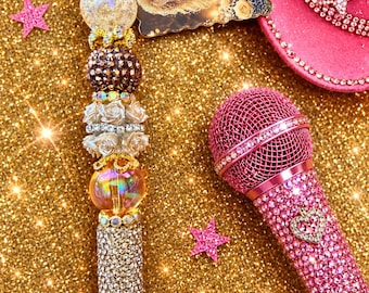 May include: A pink microphone covered in rhinestones with a heart charm, a beaded pen featuring a portrait tag labeled Remembering Dolly Parton 2026, and a pink cowboy hat with rhinestone trim and a star accent, all on a gold glitter surface.