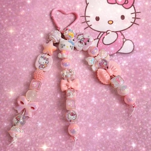 May include: Pink beaded keychain featuring Hello Kitty charms, bows, and star-shaped beads. The keychain includes a heart-shaped clasp and is set against a sparkly pink background. A Hello Kitty illustration is also present.