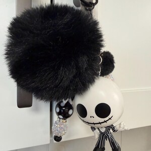 May include: A black fluffy pom-pom keyring with a decorative beaded strand and a Jack Skellington figurine. The figurine is white with black eyes and a stitched smile, wearing a pinstripe suit.