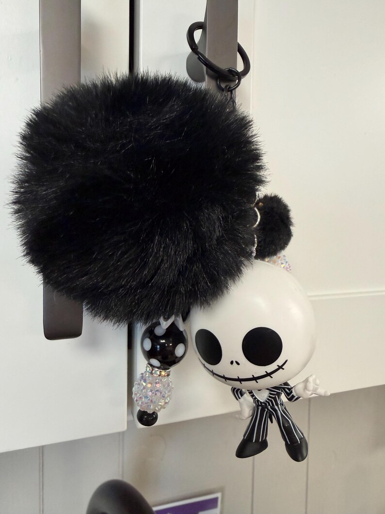 May include: A black fluffy pom-pom keyring with a decorative beaded strand and a Jack Skellington figurine. The figurine is white with black eyes and a stitched smile, wearing a pinstripe suit.