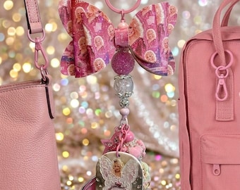 May include: A pink keychain featuring a large bow with a repeating Dolly Parton print, a strand of glittery and clear beads, and a rectangular pendant displaying an illustration of a fairy with wings. The keychain hangs from a pink metal ring.