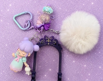 May include: A whimsical keychain featuring a purple-haired doll, a decorative arch, and a fluffy white pom-pom. The keychain includes a heart-shaped clasp and a small white bunny figurine. The color scheme is primarily pastel purple and white.