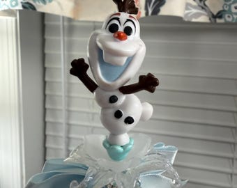 May include: A whimsical Olaf figurine, featuring a smiling face, brown twig hair, and outstretched arms. The figure is atop a decorative base with blue accents, ribbons, and sparkling embellishments. The background includes a floral curtain and a window blind.