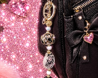 May include: A black leather backpack featuring a gold-toned metal zipper and a decorative bow. A beaded bag charm hangs from the top, consisting of gold, white, and black beads, including a crystal-encrusted sphere and a small pink heart charm.