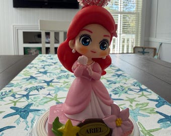 May include: A pink figurine of Ariel from The Little Mermaid, with red hair and a pink dress. The figurine is on a base with the name "Ariel" and floral accents. A decorative topper with pink and green embellishments sits above Ariel's head.