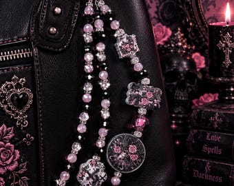 May include: A black handbag featuring a pink and black beaded charm accessory. The charm includes faceted pink and black beads, silver-toned spacers, and decorative charms shaped like a cross, a skull, and floral patterns with pink accents.