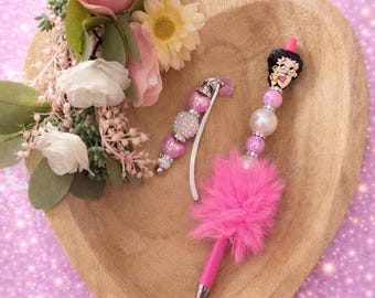 May include: A pink ballpoint pen featuring a Betty Boop character topper, a fluffy pink pom-pom, and a series of decorative pink and white beads. Beside it lies a matching beaded charm with a small pink star accent.