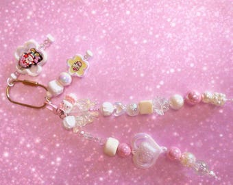 May include: A pink beaded keychain with a gold-colored clasp. The keychain features white flower-shaped charms with cartoon characters, along with various beads in white, pink, and clear colors. The background is a sparkly pink.