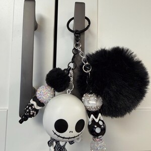May include: A novelty keychain featuring a Jack Skellington figure, black pom-pom, and decorative beads. The white Jack Skellington figure has black eyes and mouth, and wears a black and white striped suit. The keychain is attached to a black keyring.
