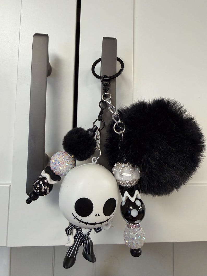 May include: A novelty keychain featuring a Jack Skellington figure, black pom-pom, and decorative beads. The white Jack Skellington figure has black eyes and mouth, and wears a black and white striped suit. The keychain is attached to a black keyring.