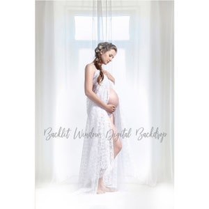 May include: A pregnant woman in a white lace dress stands in front of a window with white curtains. The woman is holding her belly and looking down. The window is backlit, creating a soft glow.