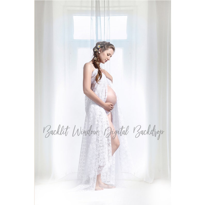 May include: A pregnant woman in a white lace dress stands in front of a window with white curtains. The woman is holding her belly and looking down. The window is backlit, creating a soft glow.