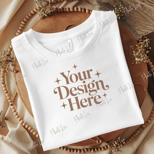 Può includere: Una t-shirt bianca a maniche corte con la scritta marrone Your Design Here, circondata da piccole icone a forma di stella a quattro punte. La maglietta ha un girocollo ed è disposta in piano, mostrando la stampa grafica centrata sul davanti.