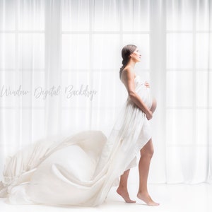 May include: A pregnant woman in a white flowing dress stands in front of a window with white curtains. The light from the window is shining through the fabric of the dress, creating a soft, ethereal glow.