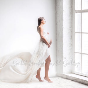 Puede incluir: Una mujer embarazada con un vestido blanco fluido se encuentra de pie frente a una gran ventana. El vestido ondea con el viento y la mujer mira hacia afuera.