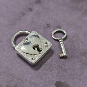 May include: A silver-colored heart-shaped padlock with a matching key. The padlock has a square base with a heart-shaped center and decorative studs. The key is small and simple, with a circular handle.