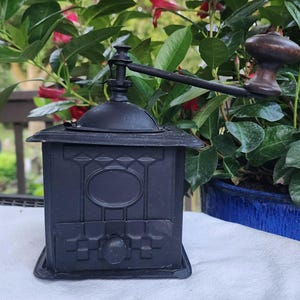 May include: A matte black coffee grinder featuring a square body with embossed geometric detailing, a central oval panel, and a small drawer with a round knob. A long metal handle extends from the top, topped with a dark brown wooden knob.