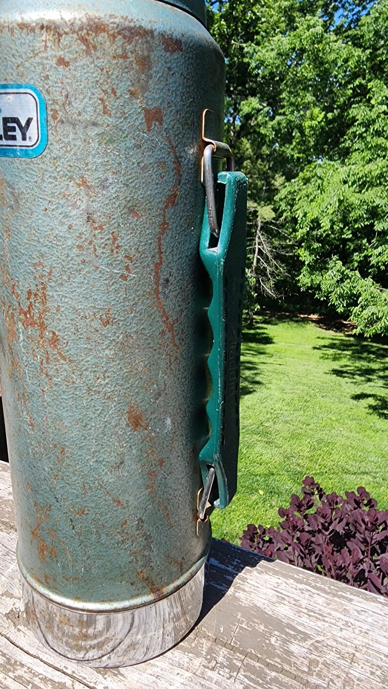 May include: Vintage Stanley thermos in a weathered teal finish with a green handle. The metal exterior shows signs of age with rust-colored spots. The thermos has a silver base and a blue and white label.