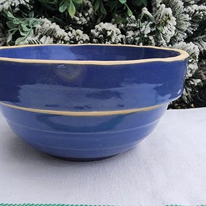 May include: A blue ceramic mixing bowl with a light beige rim and a light beige band around the middle. The bowl is sitting on a white cloth with a green and white striped edge. The background includes greenery.
