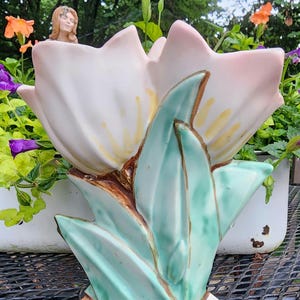 May include: A ceramic flower vase in the shape of a large, stylized flower. The petals are a soft pink with yellow accents, and the leaves are a light turquoise. The base is a warm brown color. The vase is sitting on a black metal surface.