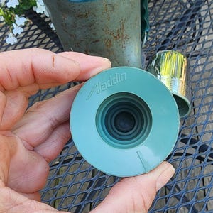 May include: A vintage Aladdin Stanley thermos with a teal lid and a matching teal stopper. The thermos has a green handle and a metal cup. The Aladdin Stanley logo is visible on the thermos.