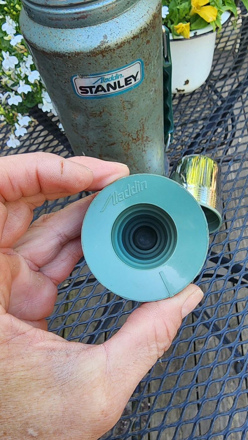 May include: A vintage Aladdin Stanley thermos with a teal lid and a matching teal stopper. The thermos has a green handle and a metal cup. The Aladdin Stanley logo is visible on the thermos.
