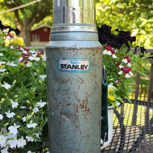 May include: Vintage Aladdin Stanley thermos in a weathered teal finish. The thermos has a shiny silver lid and a green handle. The Aladdin Stanley logo is visible on the front. The thermos is sitting on a black metal table.