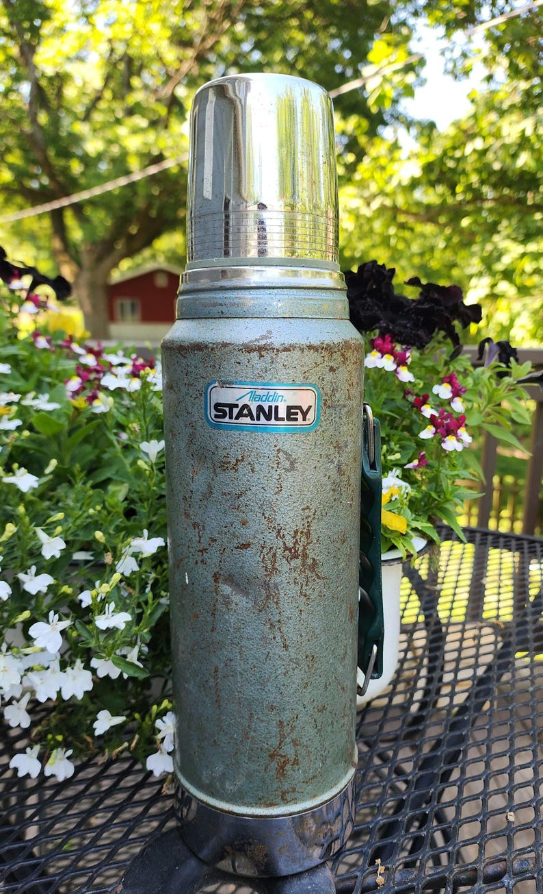 May include: Vintage Aladdin Stanley thermos in a weathered teal finish. The thermos has a shiny silver lid and a green handle. The Aladdin Stanley logo is visible on the front. The thermos is sitting on a black metal table.