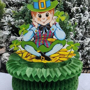 May include: A St. Patrick's Day decoration featuring a leprechaun figure sitting on a pot of gold. The leprechaun wears a green hat, vest, and pants. The pot is made of green honeycomb paper.