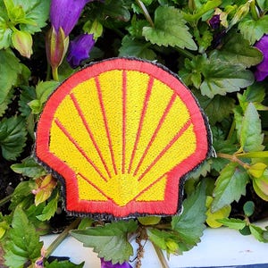 May include: An embroidered patch in the shape of a scallop shell, featuring a bright yellow center with radiating red lines and a thick red border, finished with a black stitched edge, showcasing a classic shell logo design.