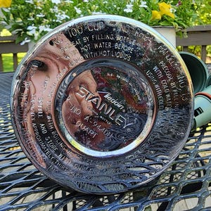 May include: A silver Stanley Aladdin quart bottle with a green cap. The bottle has text around the rim, including "100 CUP" and "STANLEY". The cap is detached and lying next to the bottle on a metal table.