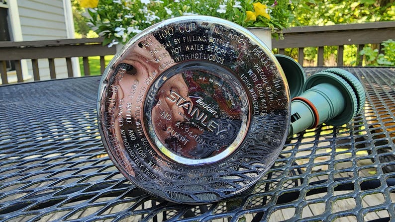 May include: A silver Stanley Aladdin quart bottle with a green cap. The bottle has text around the rim, including "100 CUP" and "STANLEY". The cap is detached and lying next to the bottle on a metal table.