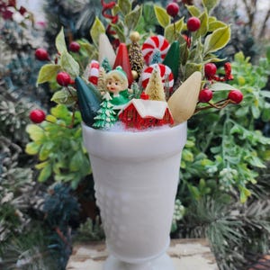 May include: A white milk glass vase filled with a miniature Christmas village scene. The scene includes a small house, a Christmas tree, a candy cane, and a small elf figurine. The vase has a textured design.