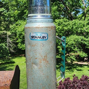 May include: Vintage Stanley thermos in a weathered teal finish with a shiny chrome lid. Features a green handle and the "Aladdin Stanley" logo. A classic insulated container for hot or cold beverages.