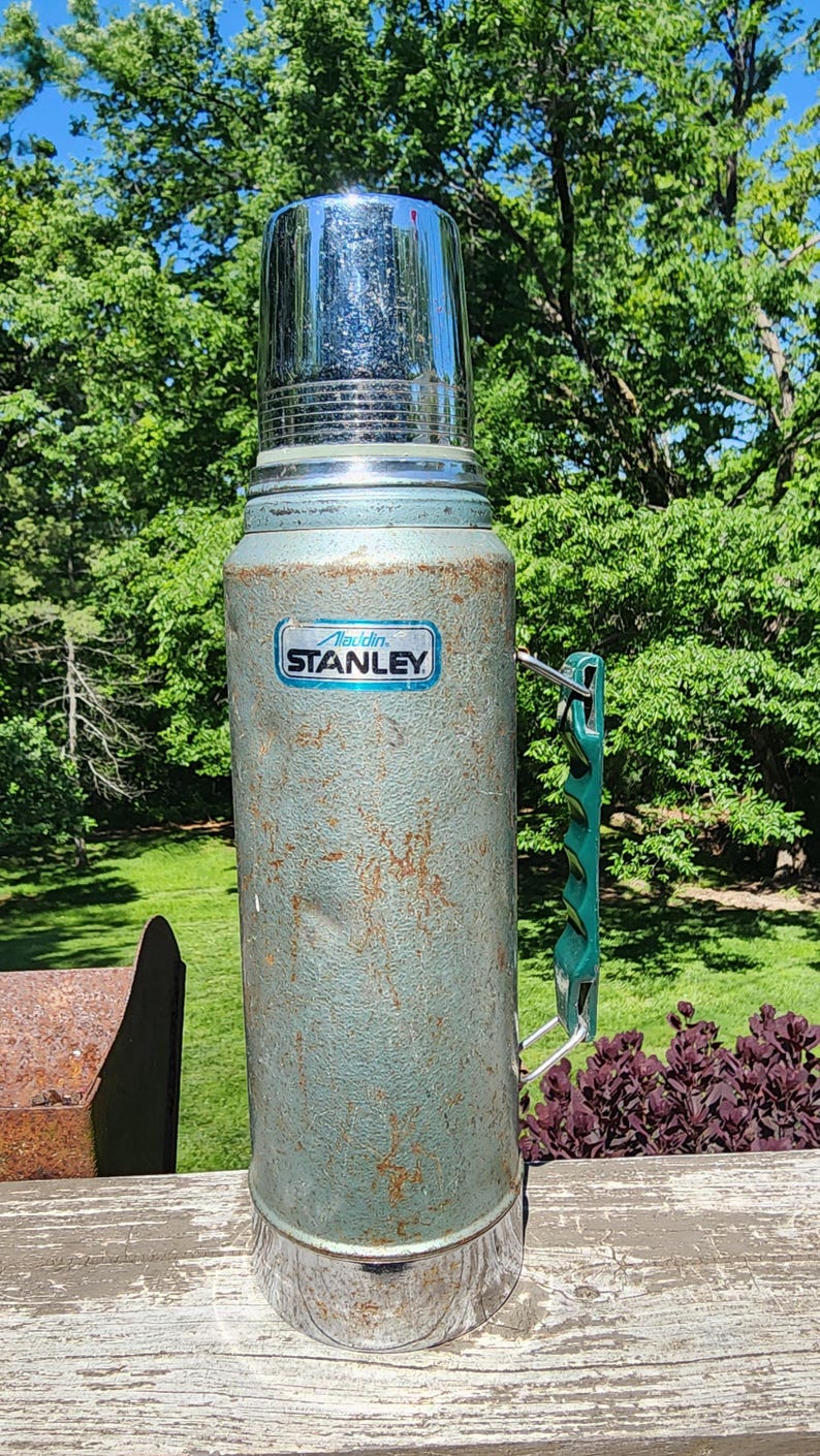 May include: Vintage Stanley thermos in a weathered teal finish with a shiny chrome lid. Features a green handle and the "Aladdin Stanley" logo. A classic insulated container for hot or cold beverages.