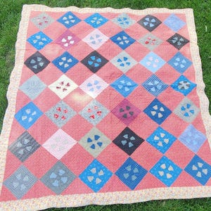 May include: A rectangular quilt featuring a repeating diamond pattern in shades of coral pink, blue, grey, and white. Each diamond contains a four-petal floral applique. The quilt is finished with a light-colored floral print fabric border.