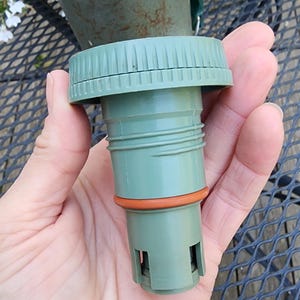 May include: A green replacement cap for a Stanley thermos. The cap has a ridged top and a cylindrical body with an orange rubber seal. The Aladdin Stanley logo is visible on the thermos in the background.