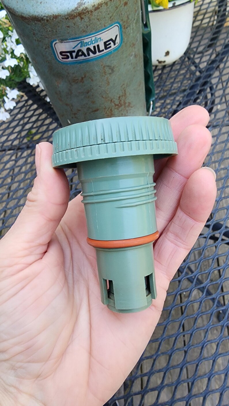 May include: A green replacement cap for a Stanley thermos. The cap has a ridged top and a cylindrical body with an orange rubber seal. The Aladdin Stanley logo is visible on the thermos in the background.