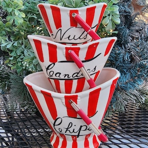 May include: Three red and white striped ceramic bowls with handles. The bowls are stacked on top of each other. The top bowl says "Nuts", the middle bowl says "Candys", and the bottom bowl says "Chips".