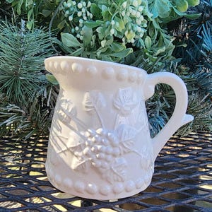 May include: White ceramic creamer with a grapevine design. The creamer has a beaded rim and handle.