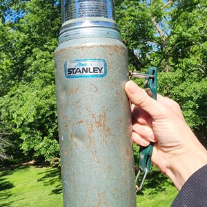 May include: Vintage Aladdin Stanley thermos in a weathered teal color with a shiny chrome top and base. The thermos has a green handle and a label that reads "Aladdin Stanley". The exterior shows signs of wear and tear.