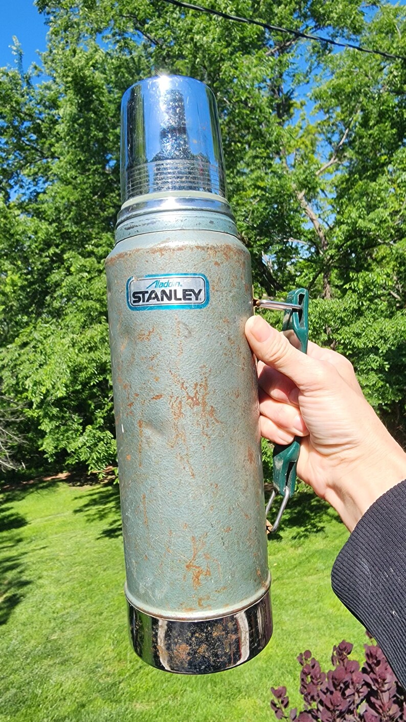 May include: Vintage Aladdin Stanley thermos in a weathered teal color with a shiny chrome top and base. The thermos has a green handle and a label that reads "Aladdin Stanley". The exterior shows signs of wear and tear.