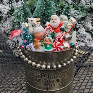 May include: A vintage Christmas display in a metal container. The arrangement includes miniature figurines of Santa Claus, a girl, and other holiday figures, along with candy canes and small Christmas trees. The container has the text "OUR SPECIAL XXX STANDARD STAMPING CO ST LOUIS".