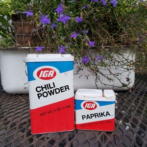 May include: Two vintage metal spice tins with red and blue labels. The larger tin is labeled "IGA Chili Powder" with a net weight of 4 ounces (113g). The smaller tin is labeled "IGA Paprika" with a net weight of 1 1/4 ounces (35g).