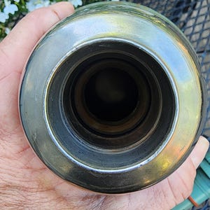 May include: A shiny, metallic bottle stopper with a circular design. The stopper has a polished, reflective surface and a series of concentric rings leading to a dark interior. The object is held in a hand, with a blurred background.