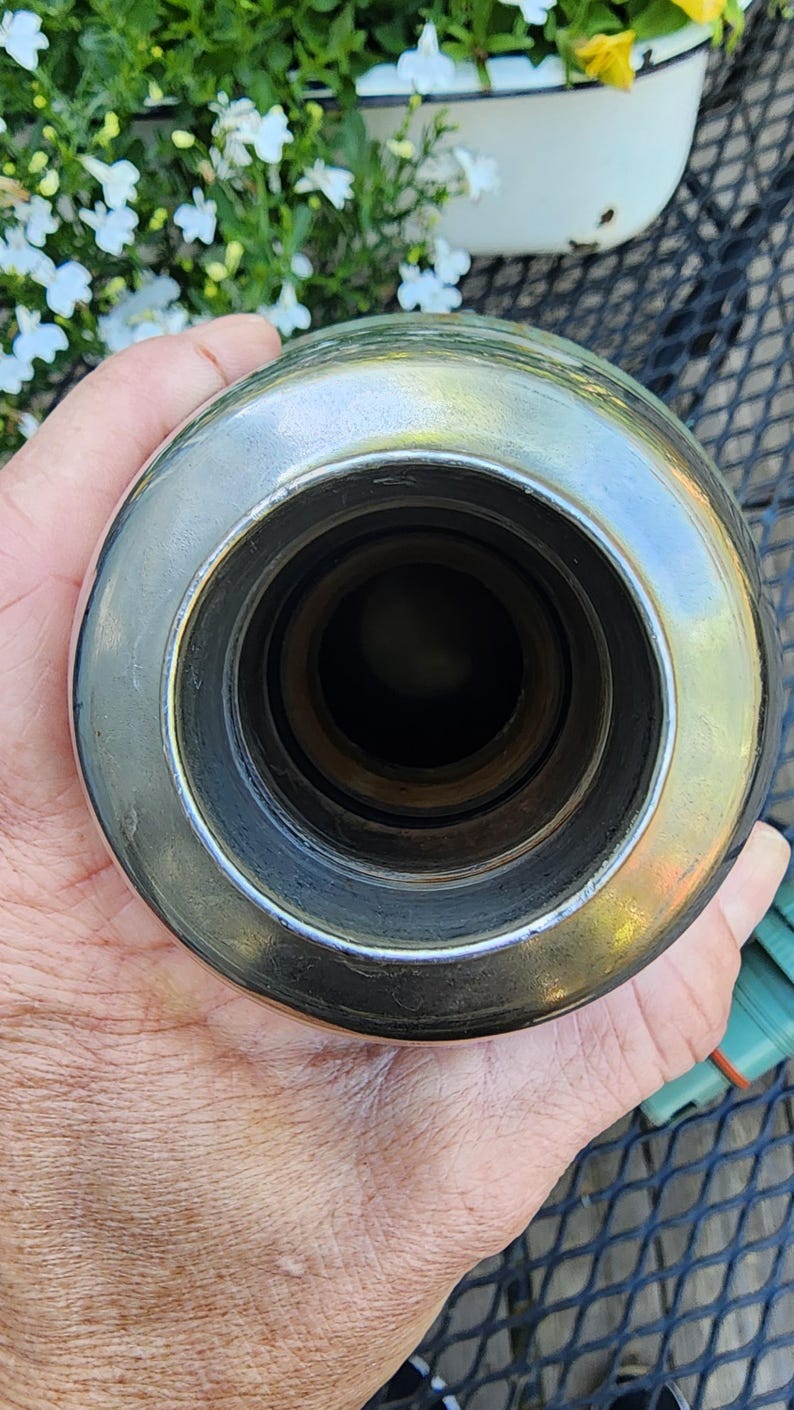 May include: A shiny, metallic bottle stopper with a circular design. The stopper has a polished, reflective surface and a series of concentric rings leading to a dark interior. The object is held in a hand, with a blurred background.