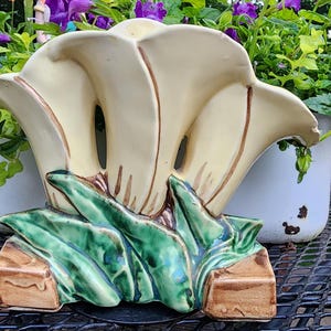 May include: A vintage ceramic flower vase in the shape of a calla lily. The vase is cream-colored with brown accents and green leaves at the base. The vase is placed on a black metal table with purple flowers in the background.